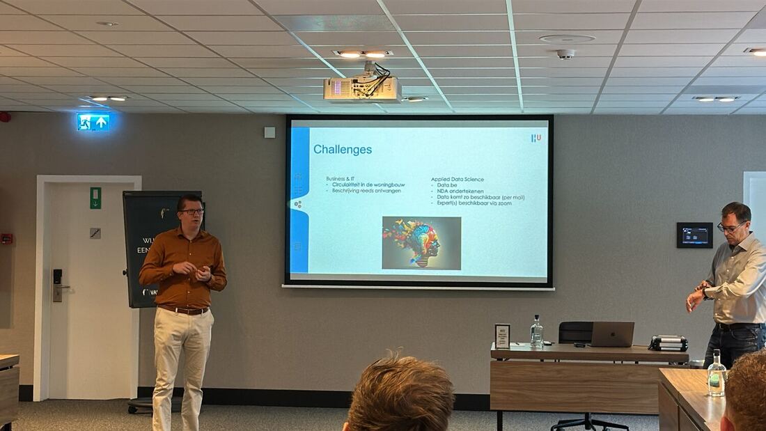 Two presenters stand beside a screen showing a slide headed Challenges, listing the Business and IT brief and the Applied Data Science brief with Data.be.