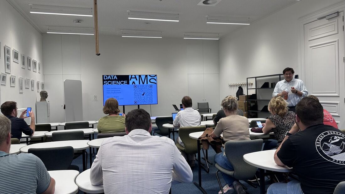 An audience seated at small tables faces a screen reading Data Science AMS, advertising a talk on data architecture, integration and deployment.