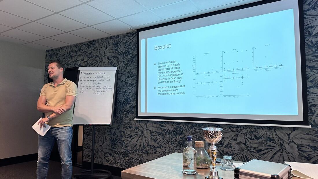 A presenter at a large screen showing a table of results, with a flip chart alongside.