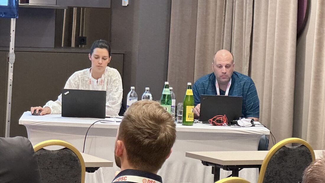 Two instructors work from laptops at a table at the side of the room during a hands-on session, a delegate seated in front of them.
