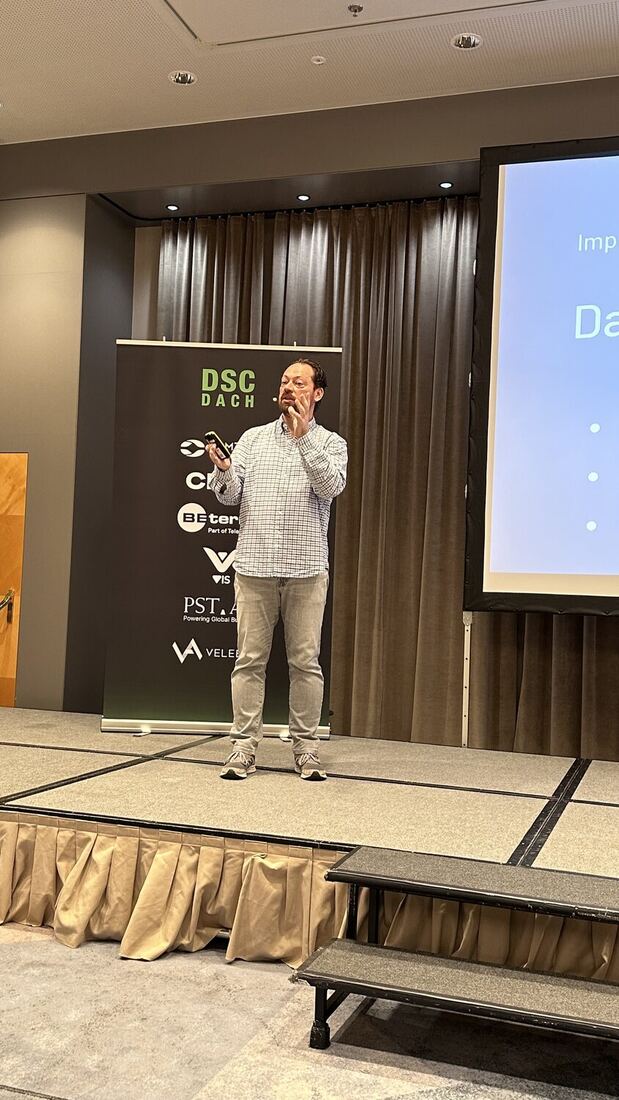A speaker with a microphone on stage in front of the screen, a DSC DACH banner behind him.