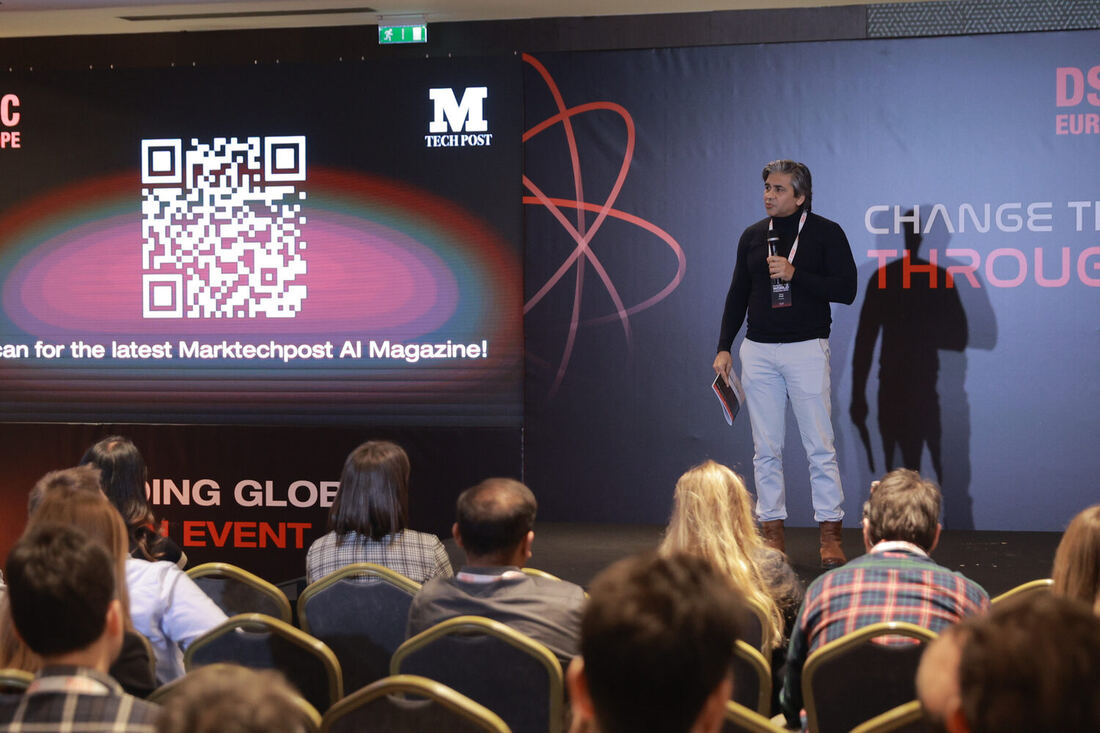 A speaker on the main stage beside a screen carrying a QR code for the Marktechpost AI magazine, the audience seen from behind
