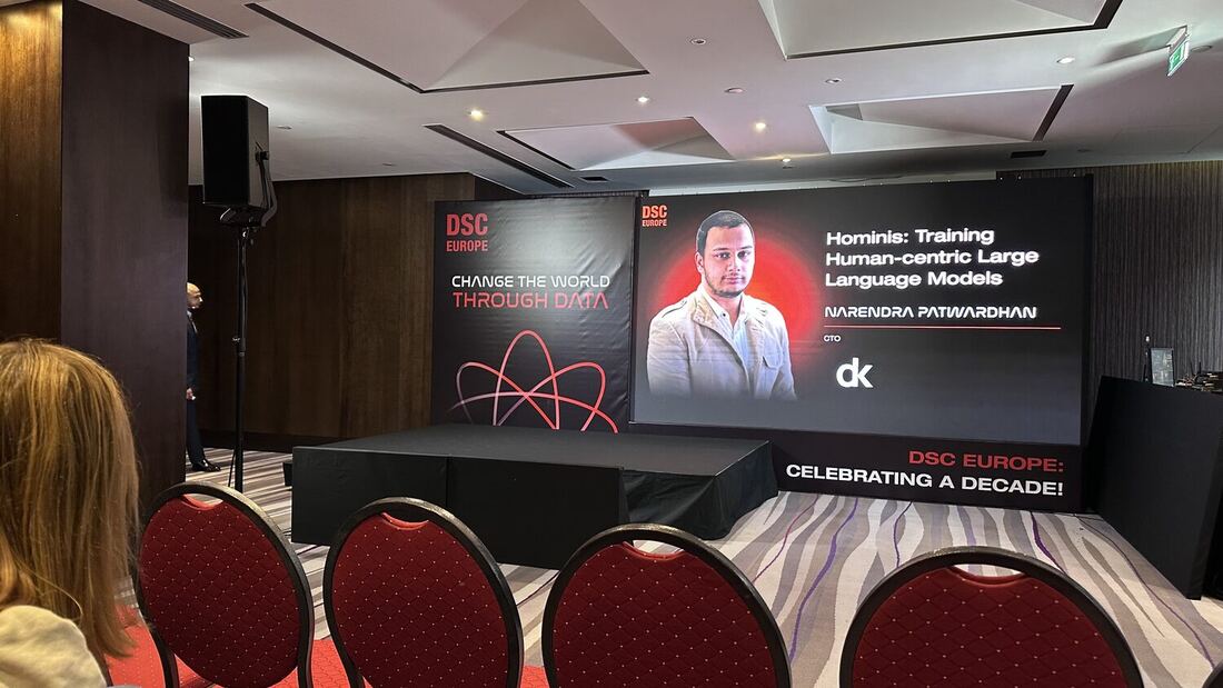 The stage screen carries the title card for Hominis: Training Human-centric Large Language Models, billed to Narendra Patwardhan, CTO, under the dk mark