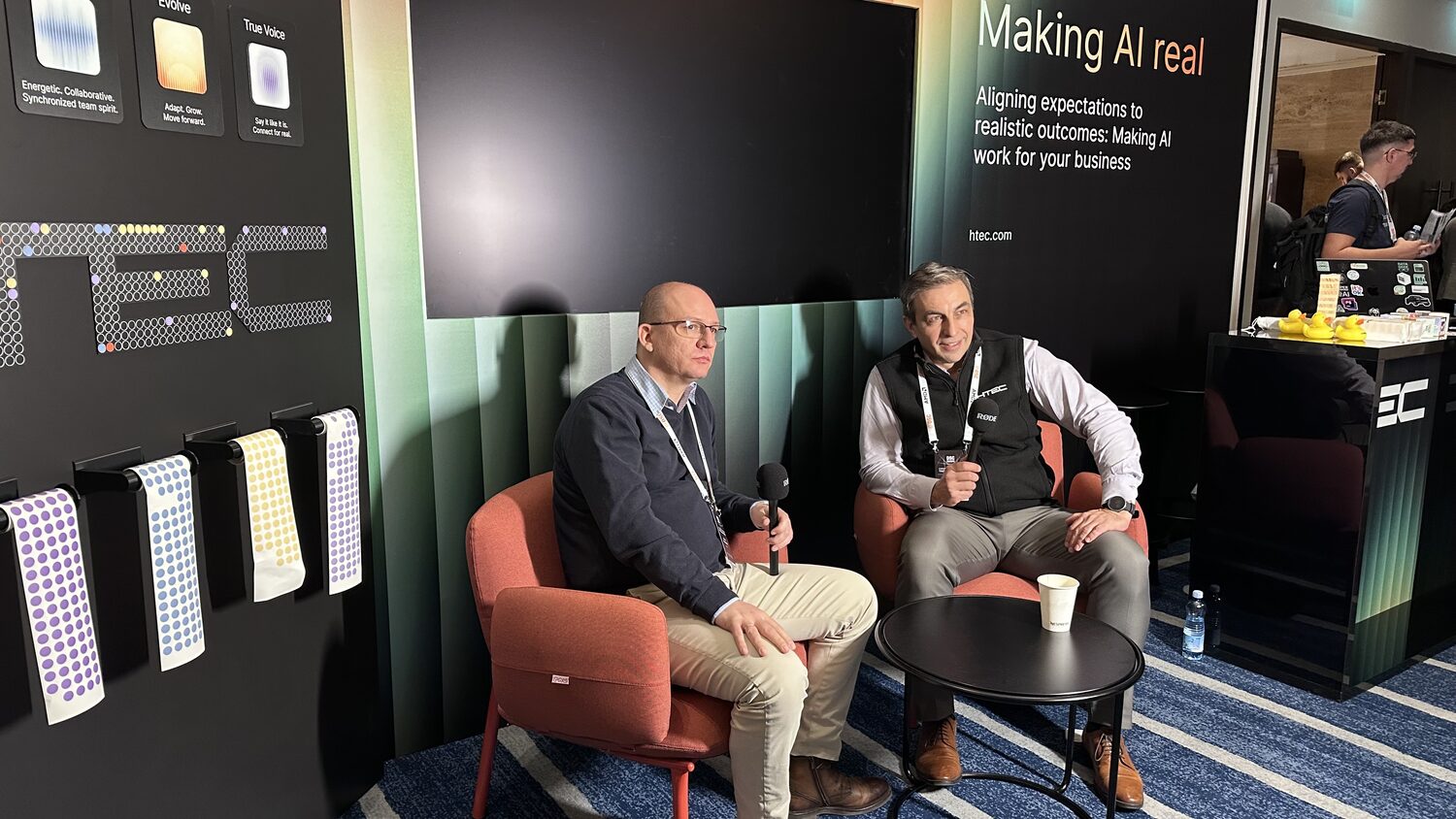 Two men recording an interview in orange armchairs at the HTEC stand, under wall copy reading Making AI real.