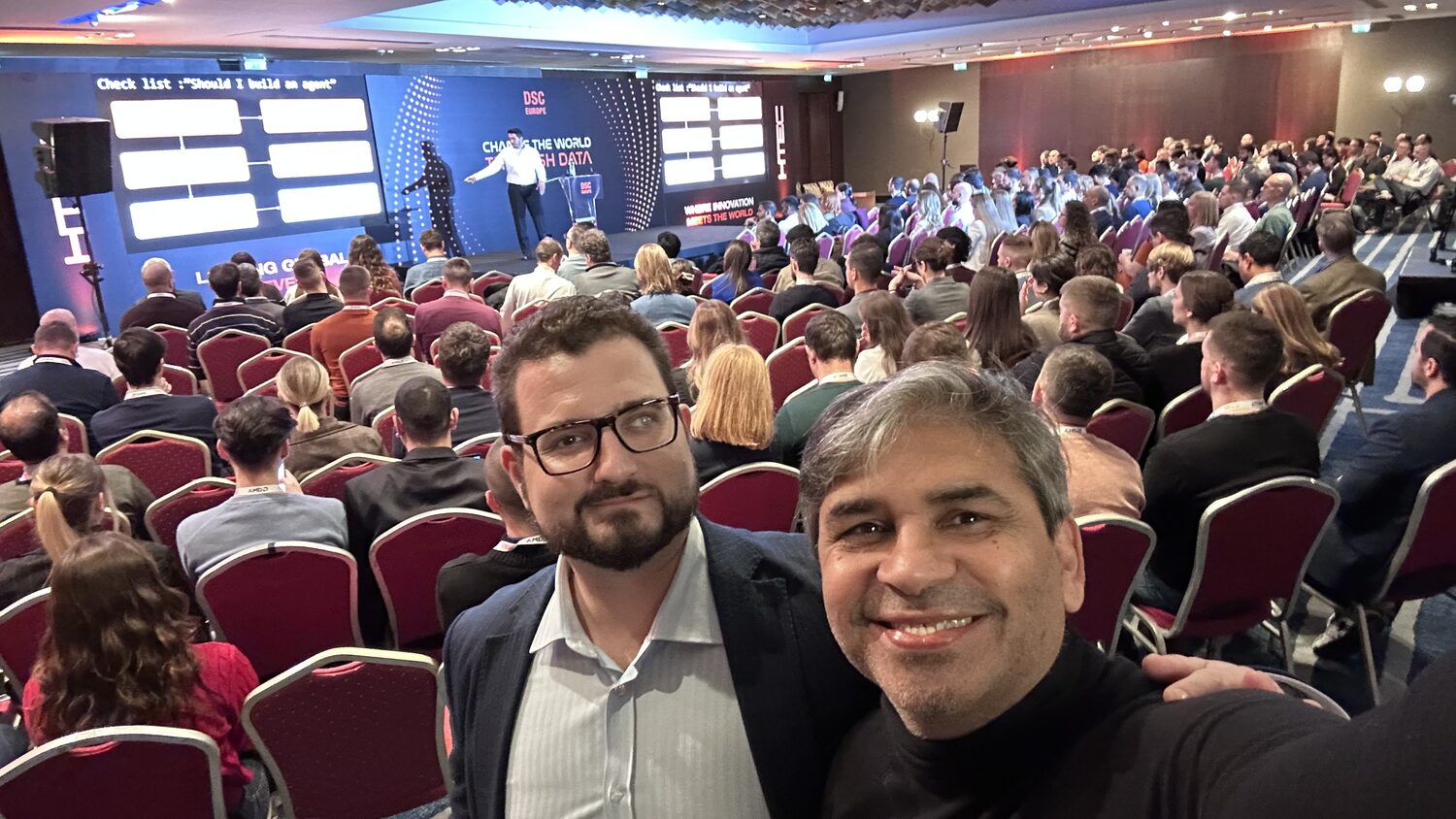 Two delegates at the back of the full main hall, the stage and slide screens visible behind them.