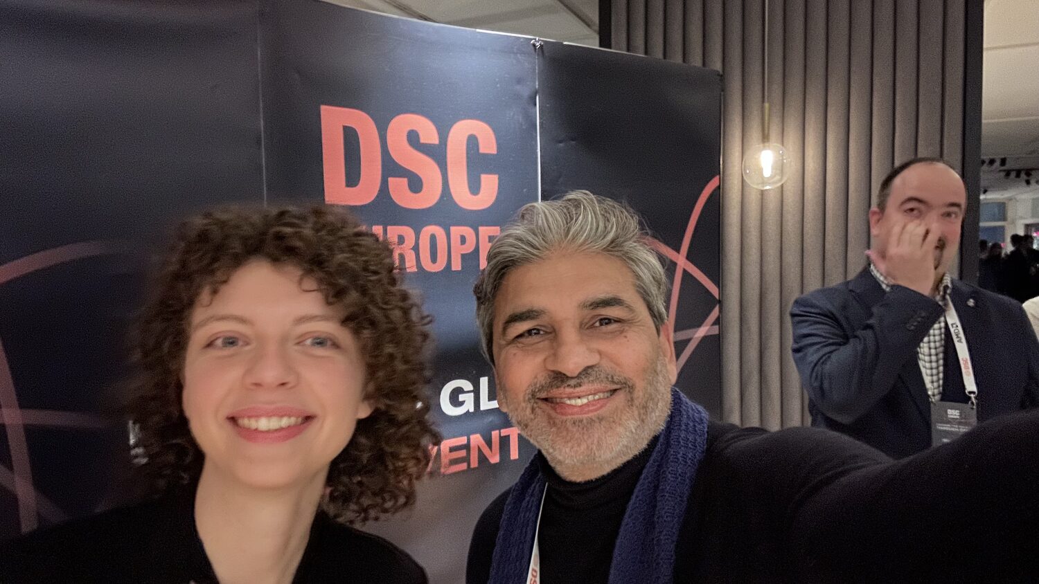 Two people in front of the DSC Europe backdrop, a delegate in a lanyard passing behind them.