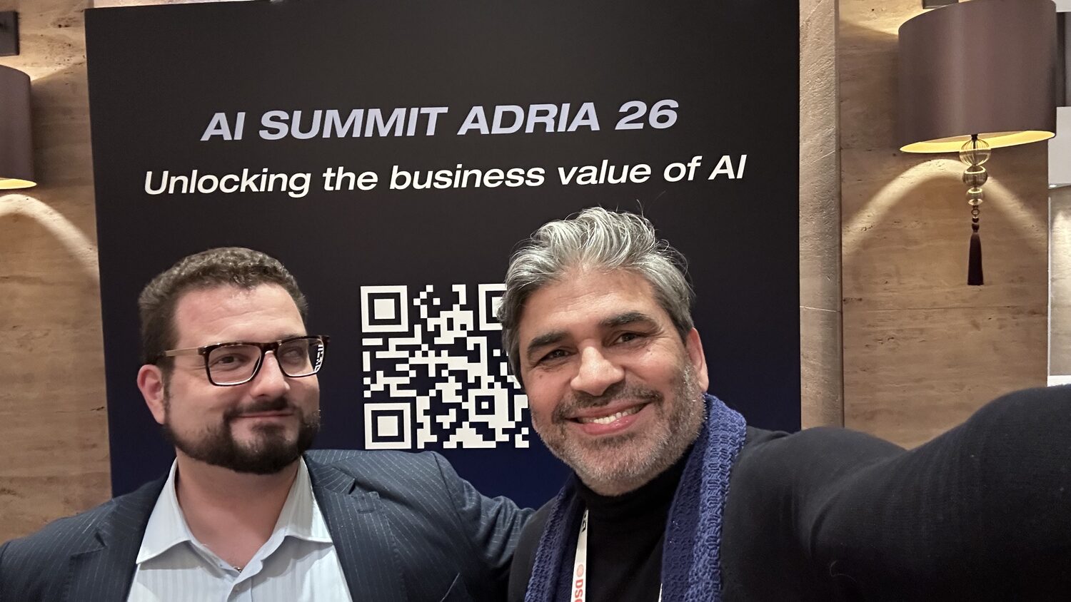 Two people in front of a black banner reading AI Summit Adria 26, Unlocking the business value of AI, with a QR code.