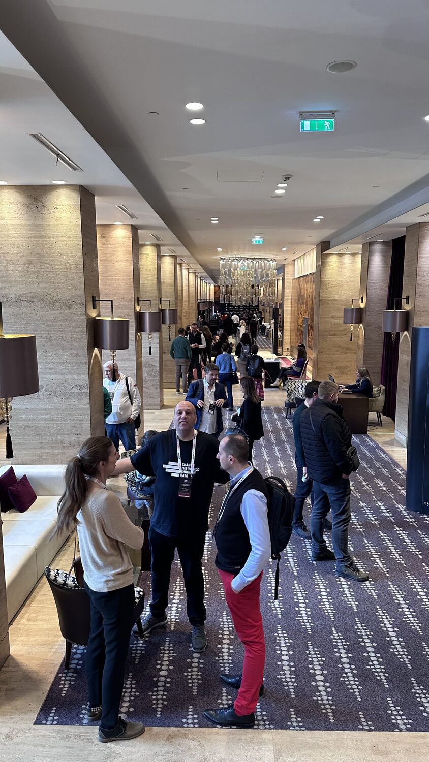 Delegates talking in the venue foyer, corridor of lamps and travertine columns running back to the chandelier.
