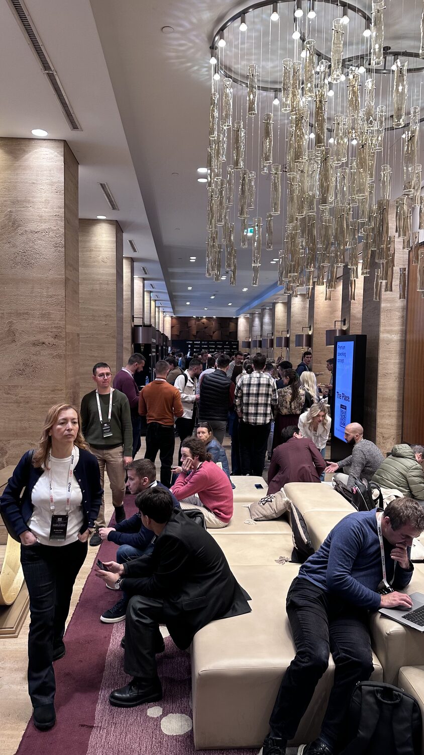 Coworking lounge under a hanging glass chandelier, delegates on long benches with laptops, a screen advertising the coworking area.