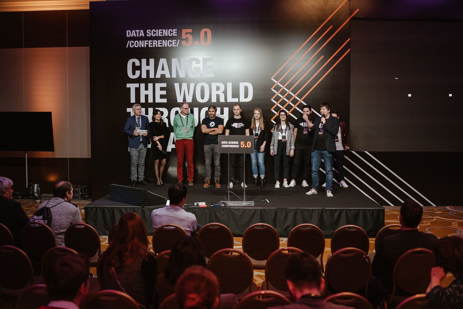 A group on the main stage at the close of a session, the conference backdrop behind and the hall full in front of them.