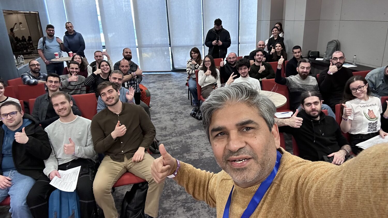 Workshop cohort of about thirty seated in red chairs, most with a thumb raised, photographed from the front of the room
