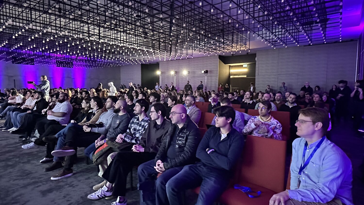 Packed conference audience in the main hall, seated under a steel ceiling grid hung with bare bulbs