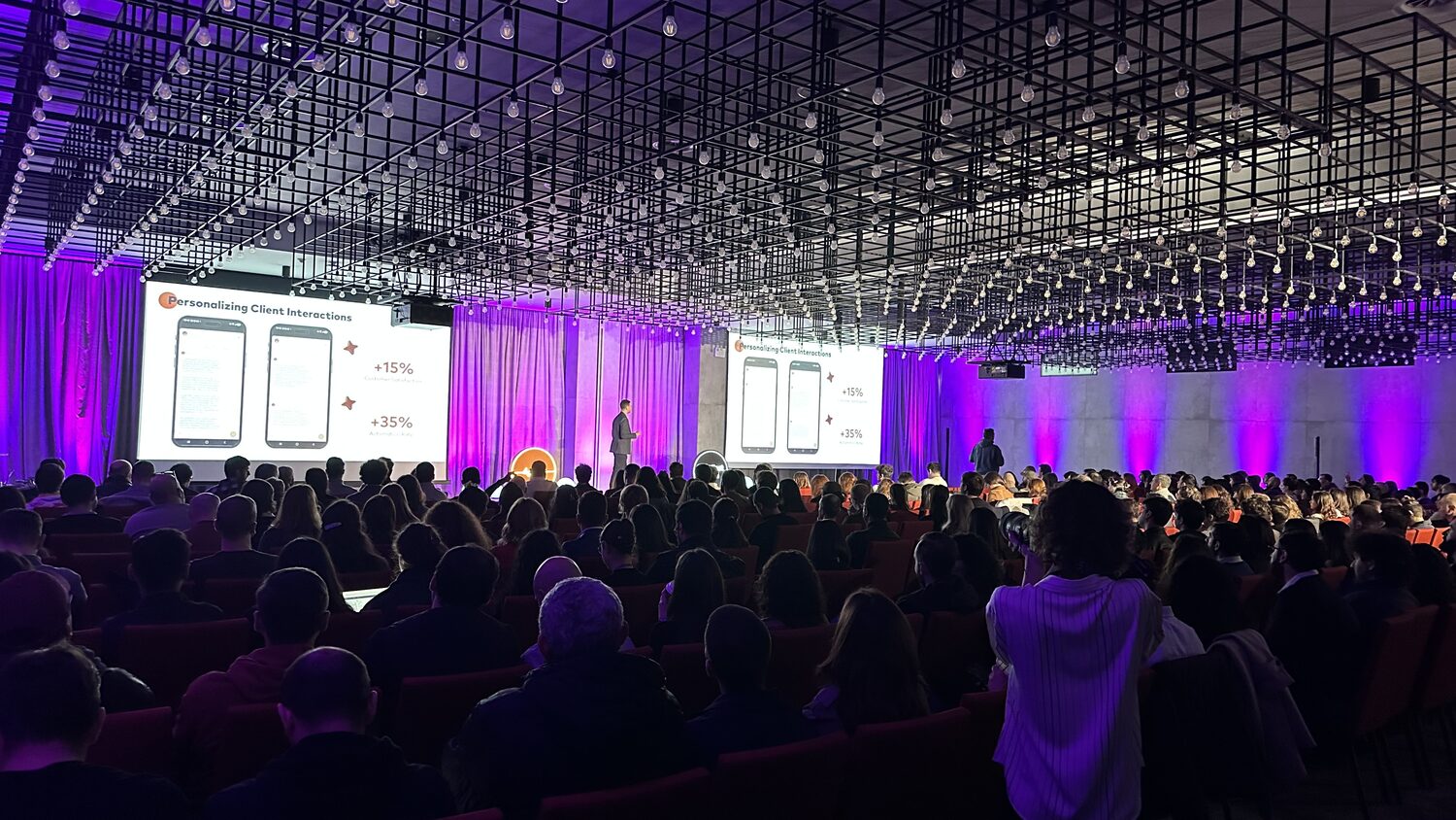 Wide view of the full main hall from the back rows, a speaker on stage and slides on two screens