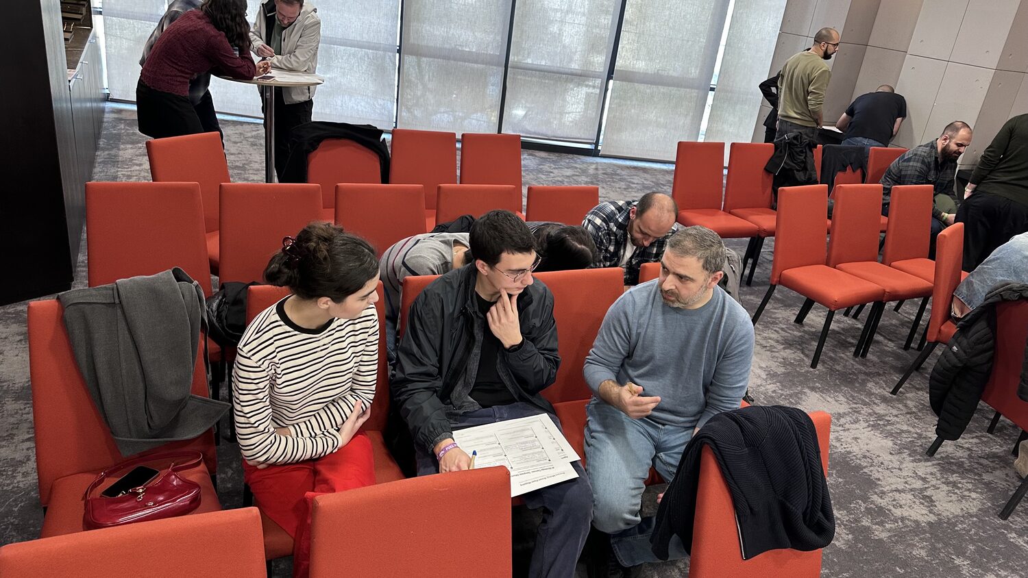 Three participants talking over a printed canvas in the red chairs, other groups working behind them
