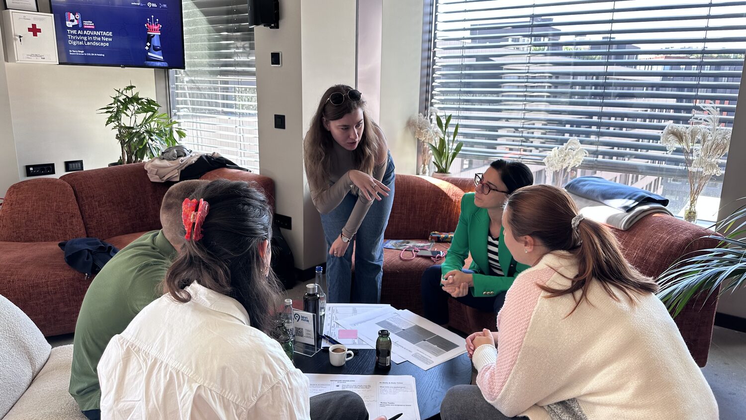 A facilitator leans in over a sofa group of five working through a worksheet, a wall screen showing the workshop title slide.