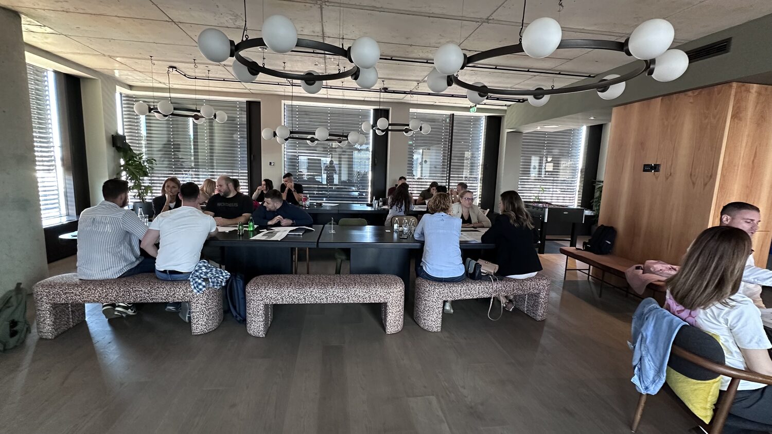 Long tables and upholstered benches across the office floor, groups talking in twos and threes.