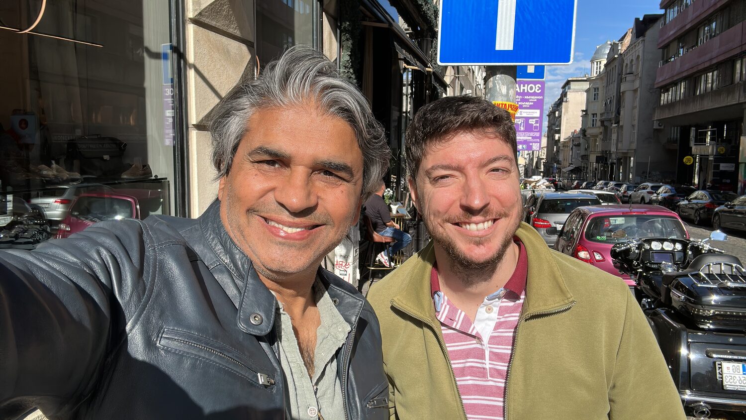 Two men smiling in a selfie on a central Belgrade street, parked cars and a blue road sign behind them.