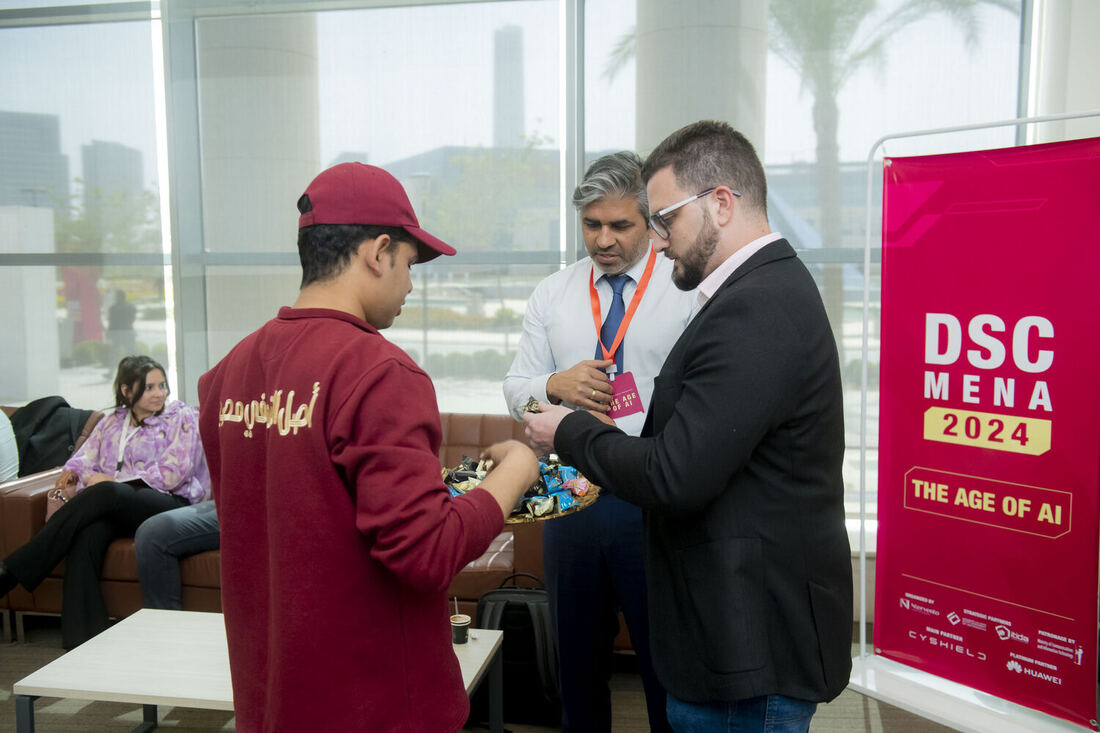 A student in a red cap speaking with two men beside a DSC MENA 2024 roll-up banner in the university foyer.