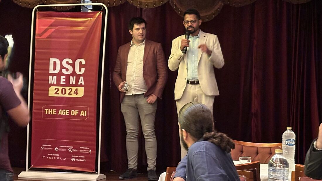 A speaker with a microphone addressing a seated group beside a DSC MENA 2024 banner at the welcome function the evening before the conference.