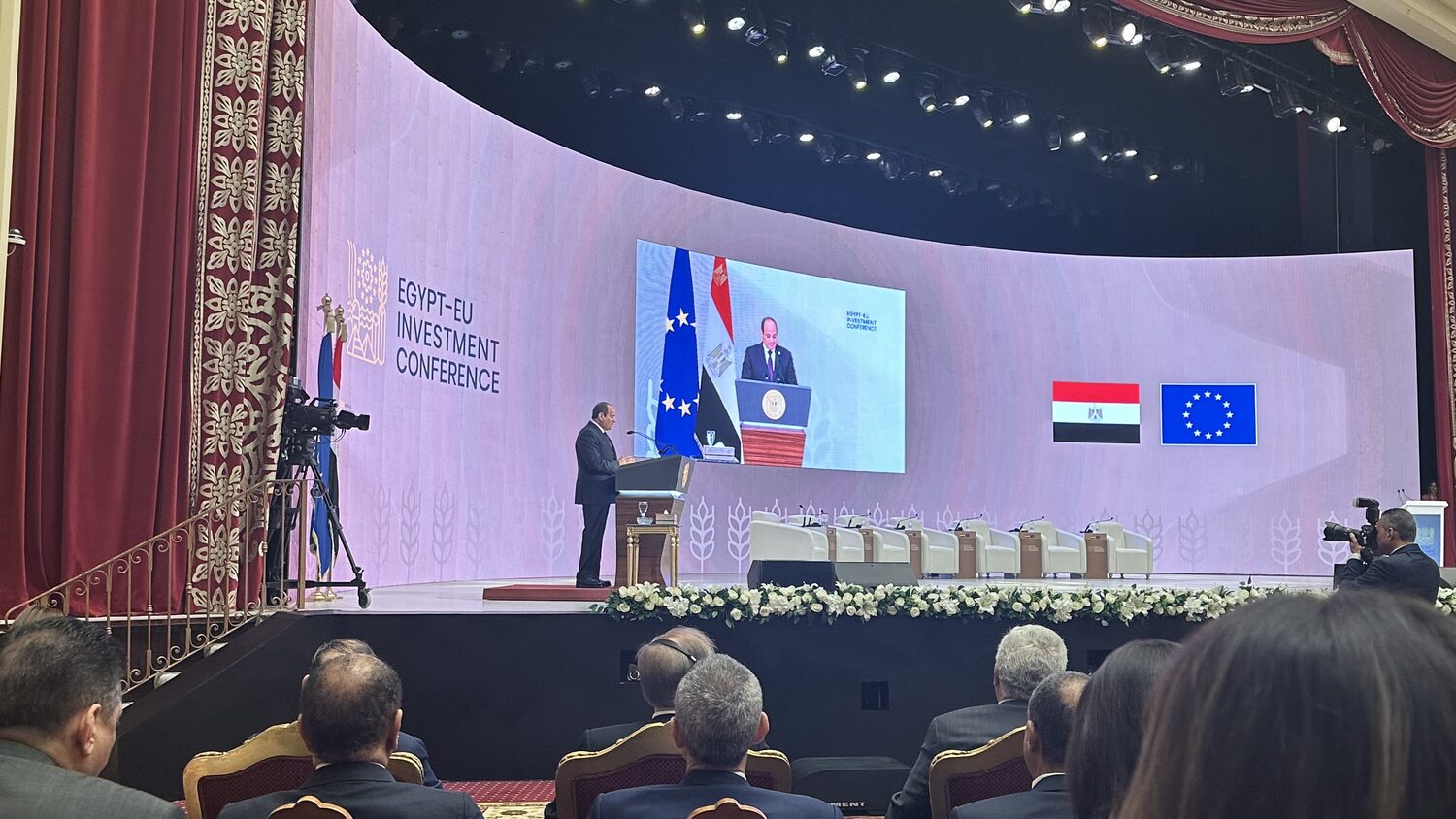 President Abdel Fattah El-Sisi speaks from the lectern at the Egypt-EU Investment Conference, relayed on the curved stage screen beside the Egyptian and EU flags, the audience seated in front.