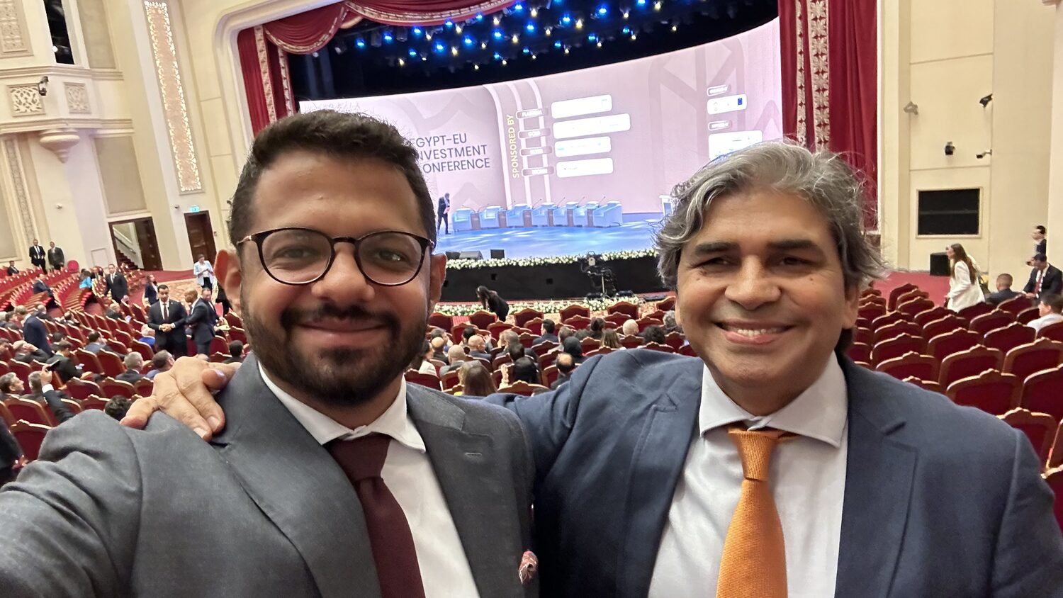 Two delegates before a session in the Al Manara theatre, the conference stage and the filling rows of red seats behind them.