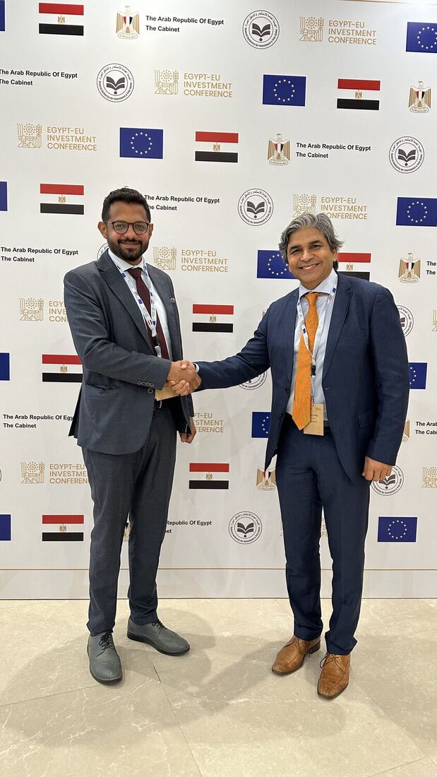 Two delegates shake hands in front of the conference wall, both wearing accreditation badges.
