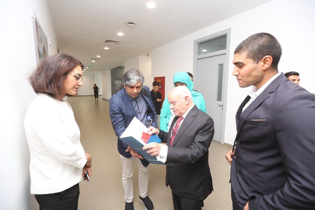 A corridor handover: a host opens a bound document to show Tarry Singh, with staff and students standing around them.