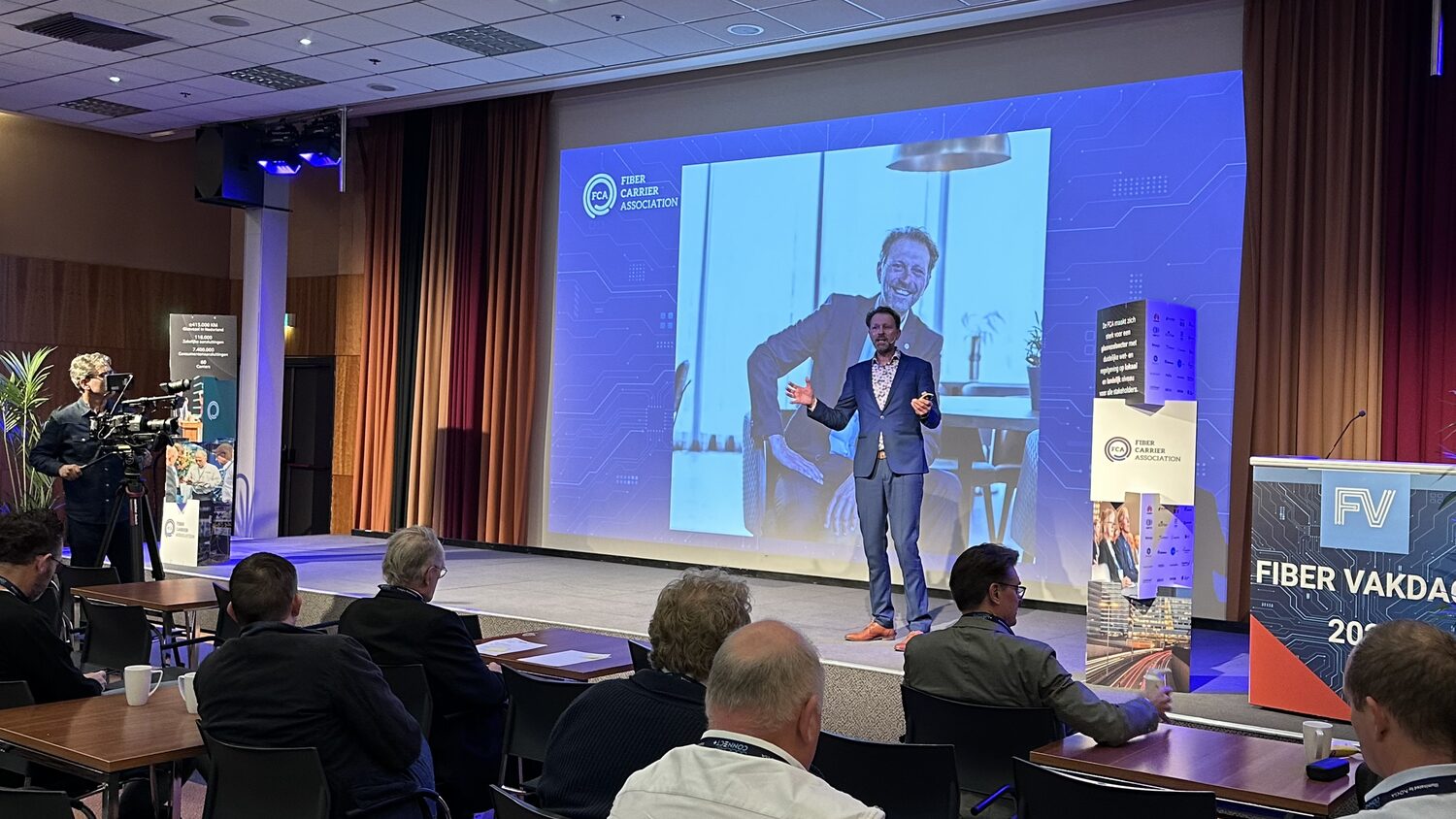 A speaker in a blue suit works the Fiber Vakdag stage in front of a large projected portrait, delegates seated at tables in the foreground and a camera operator filming from the left.