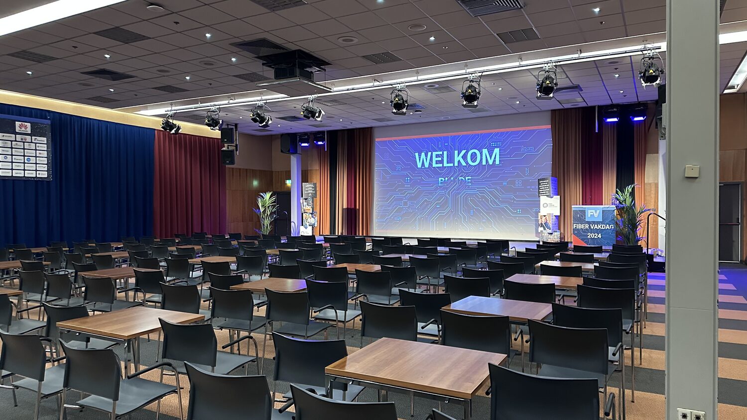The Fiber Vakdag hall laid out with tables and chairs before the first session, WELKOM on the main screen and the FIBER VAKDAG 2024 roll-up banner beside the stage.