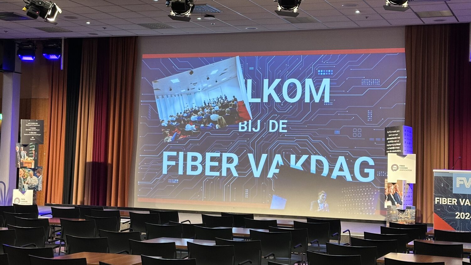 The welcome slide reading WELKOM BIJ DE FIBER VAKDAG fills the main screen, with Fiber Carrier Association display pillars standing either side of the empty stage.