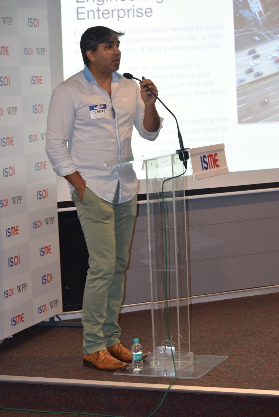 Tarry Singh presenting from a perspex lectern at ISME Tower, Lower Parel, a slide projected behind him and an ISME and ISDI branded backdrop to his left.