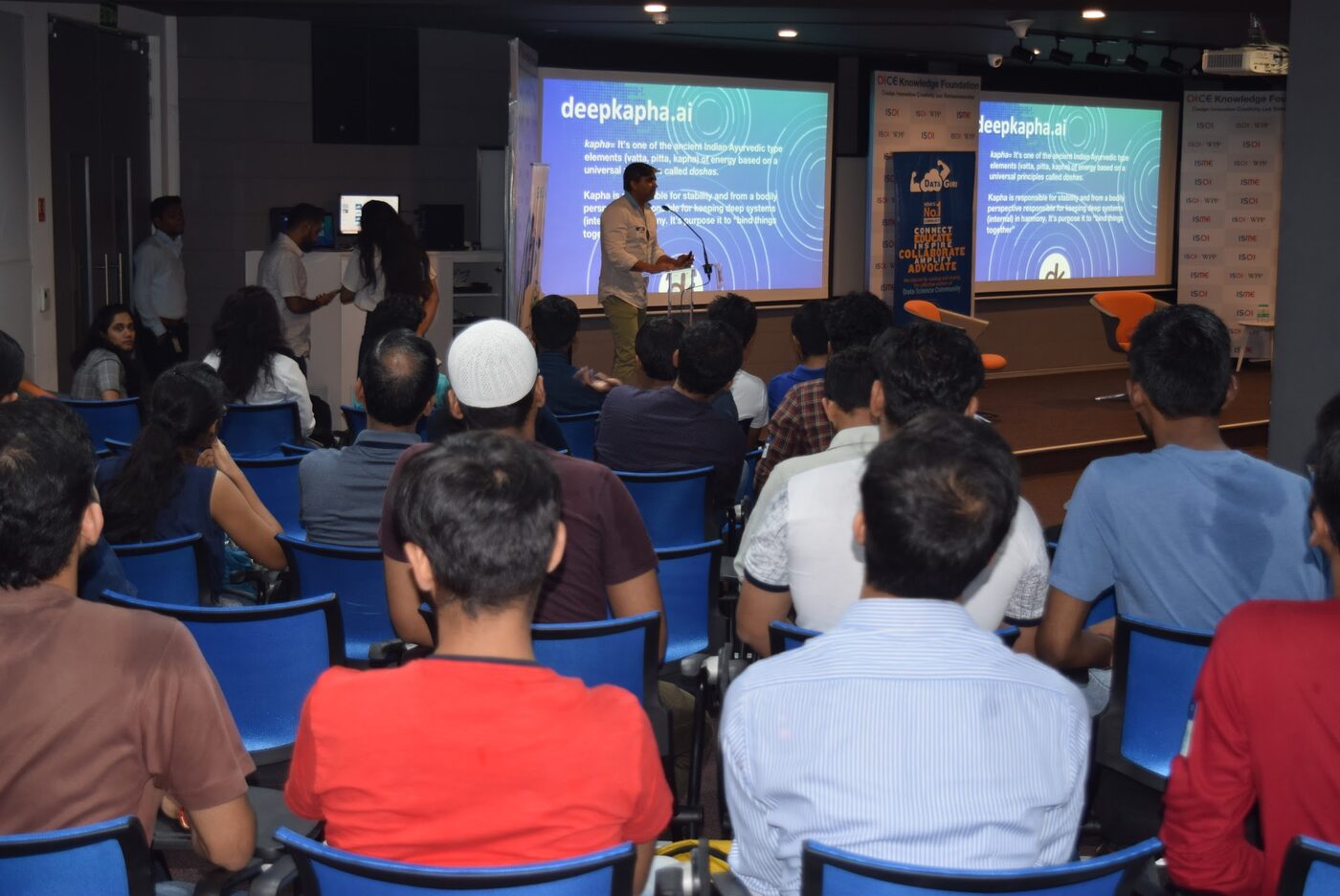 The audience seen from behind, facing twin screens carrying deepkapha.ai slides in the ISME Tower auditorium.