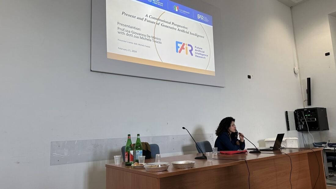 A presenter speaks into a desk microphone under the title slide for a constitutional perspective on generative artificial intelligence, carrying the FAIR programme logo.
