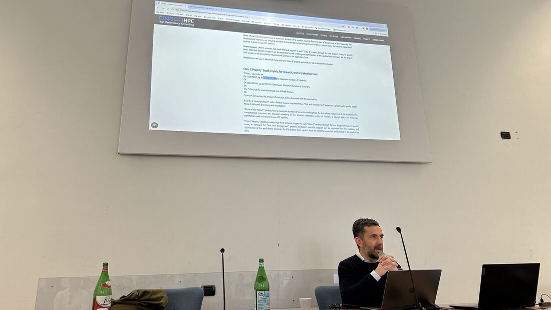 A speaker sits at the presenters' desk with two laptops while the CINECA HPC project classes page is projected behind him.