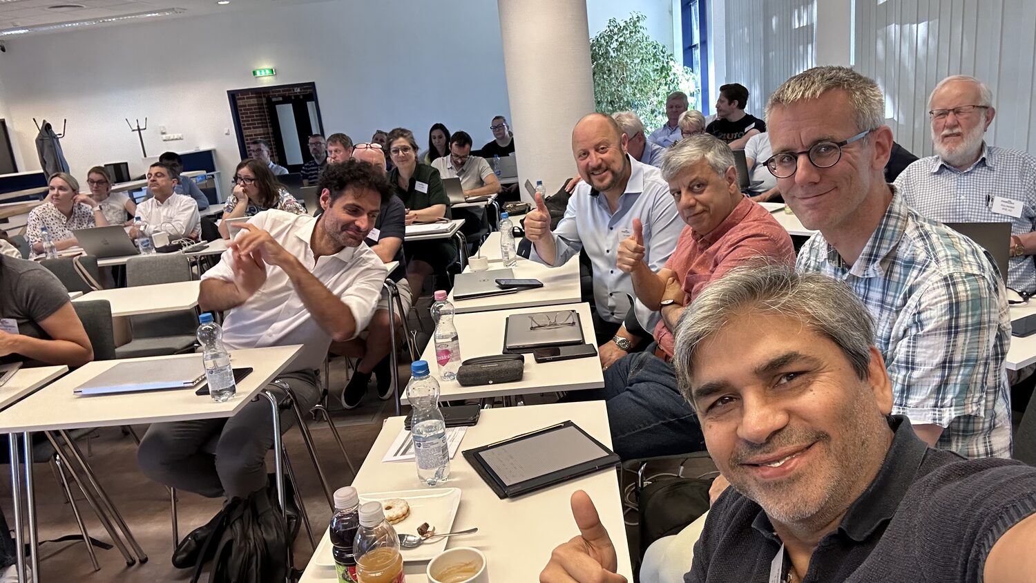 Tarry Singh with consortium colleagues in the meeting room during a break, the rest of the cohort at their desks behind.