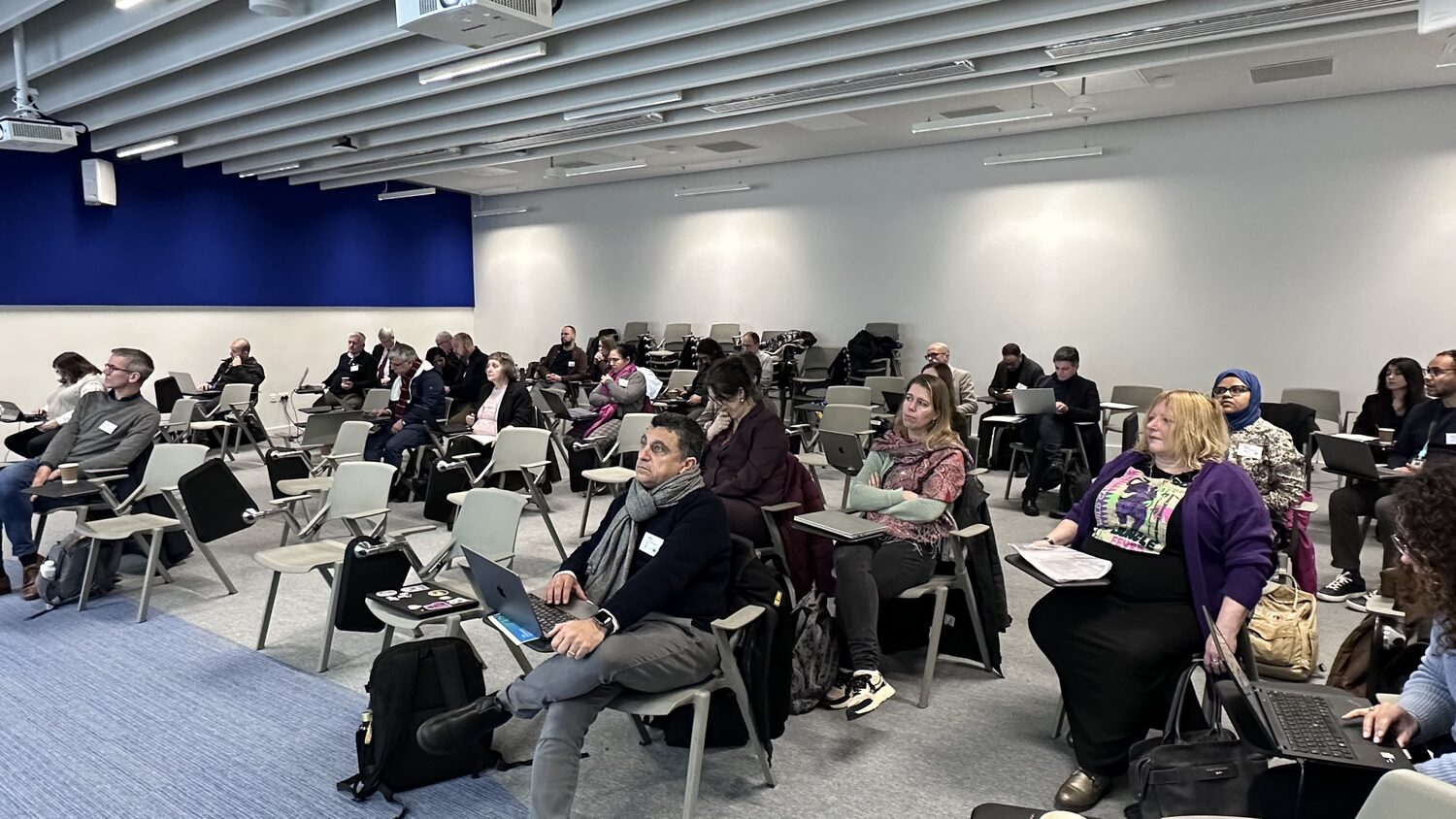 More than two dozen delegates seated with laptops and clipped name badges in a flat-floor lecture room with a deep blue wall, during the PANORAIMA consortium session in Dublin.