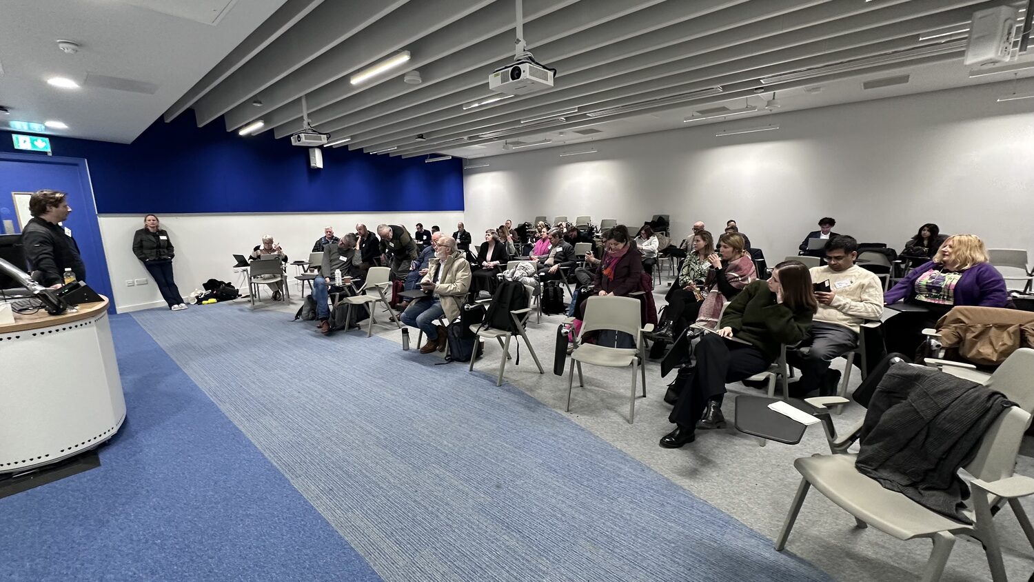 Wide view of the same lecture room late in the afternoon, lectern and microphone at the left, delegates at tablet-arm chairs across the right of the room.