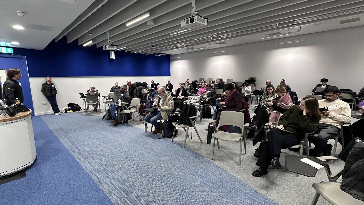 The session room from the front. Delegates work at laptops and phones, coats and bags stacked on empty chairs, as the day runs on.