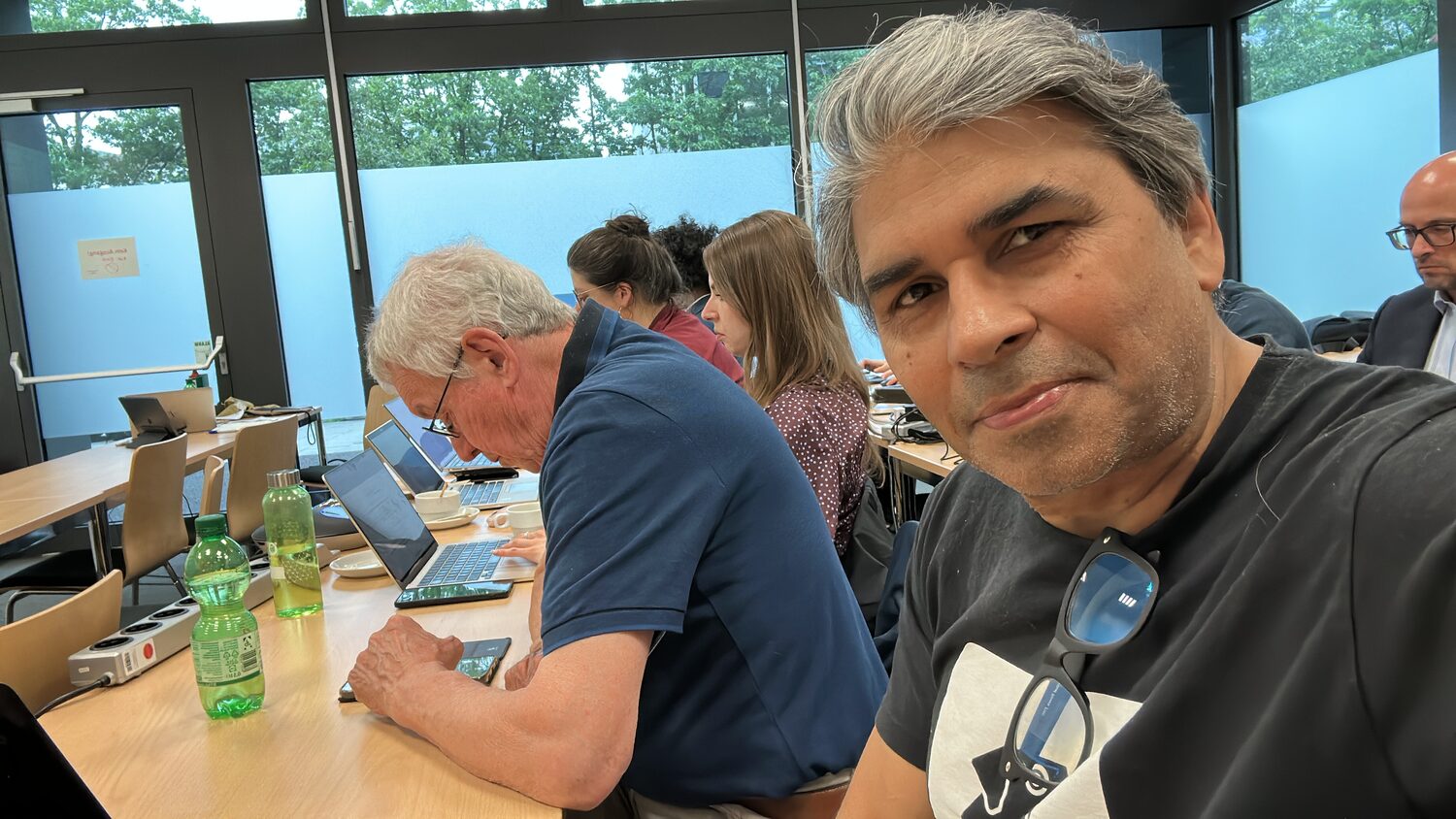 Consortium members working along a long table set against a window wall, laptops and a tablet open, cups and bottles down the middle.