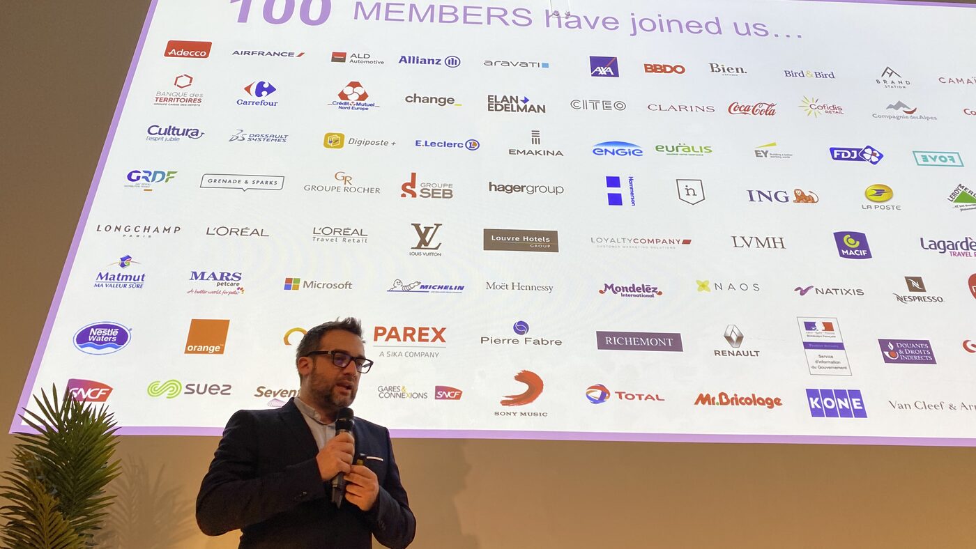 A speaker stands in front of a projected wall of a hundred HUB Institute member marks, among them Air France, Carrefour, L'Oreal, LVMH, Michelin, Renault, SNCF and Total.