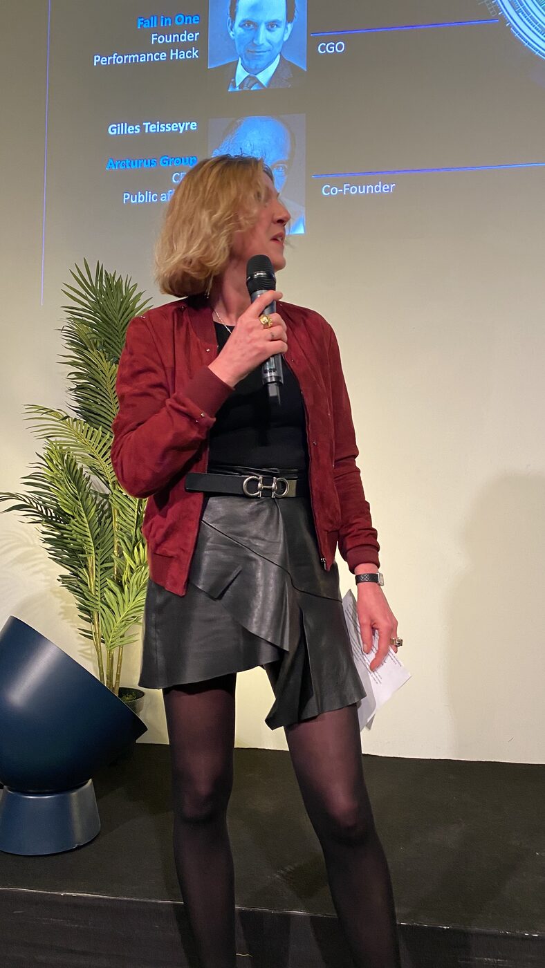 A speaker in a red jacket addresses the room, notes in one hand, the founding-team slide projected on the wall behind her.
