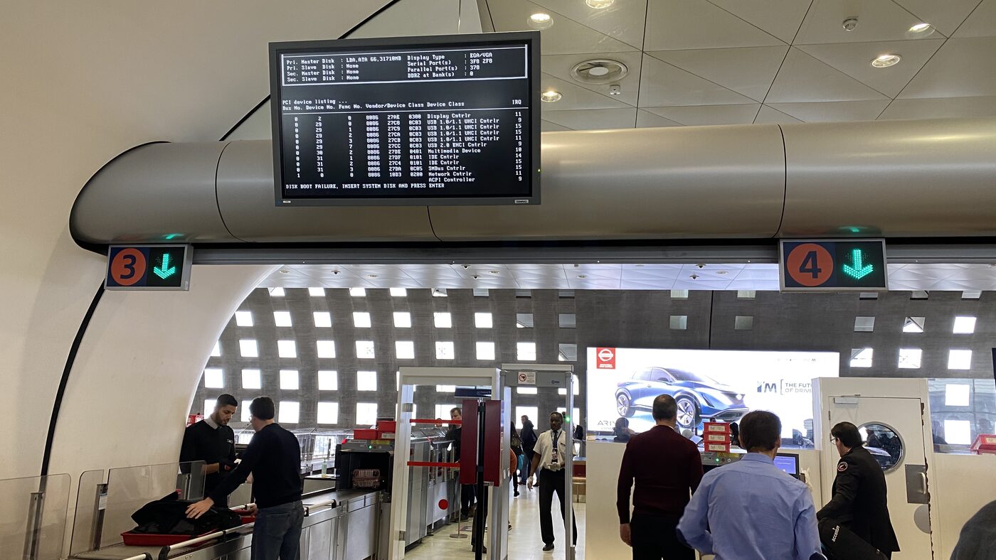 A display above security lanes three and four at Charles de Gaulle showing a BIOS device listing and the line DISK BOOT FAILURE, INSERT SYSTEM DISK AND PRESS ENTER.