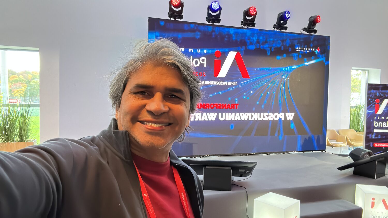 Tarry Singh in front of the AI Summit Poland 2025 stage screen, the LED wall showing the event logo, the dates 14-15 października 2025 and the Polish strapline about transformation and value.