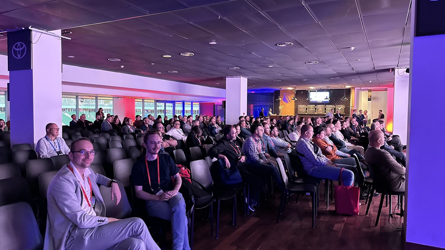 A full auditorium at the Legends Club, delegates in red lanyards seated in rows under coloured stage lighting, with stadium windows onto the pitch behind them.