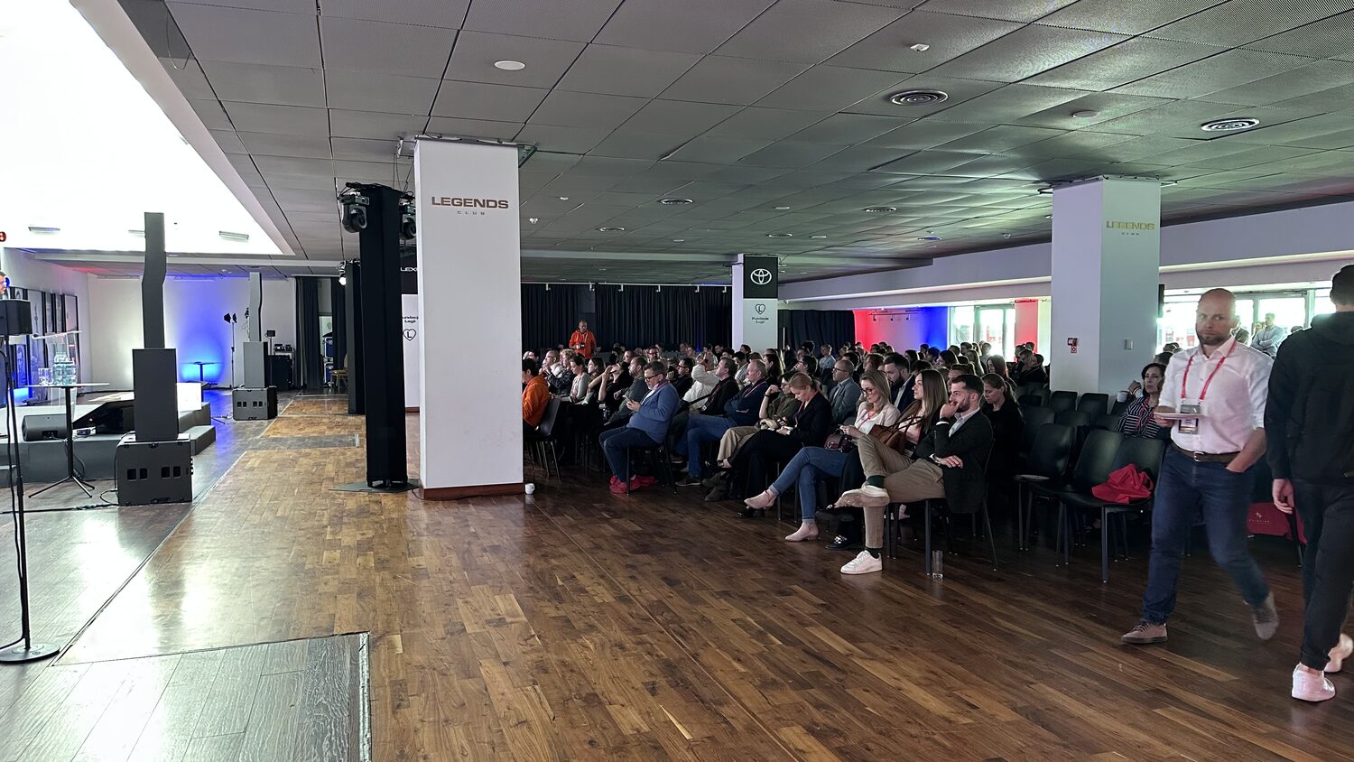 The conference floor at Legia Warsaw's stadium, pillars branded Legends Club, an audience seated to the right and the speaking platform and monitors at the left.