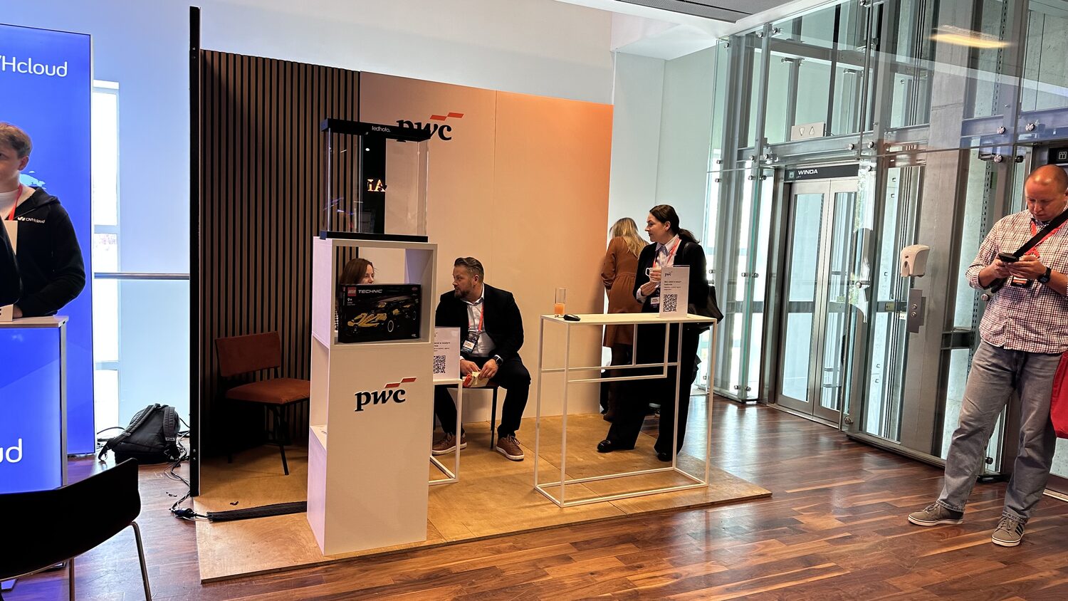 The PwC stand in the summit corridor, a wood and terracotta booth with a Lego Technic display, delegates talking beside the lifts.