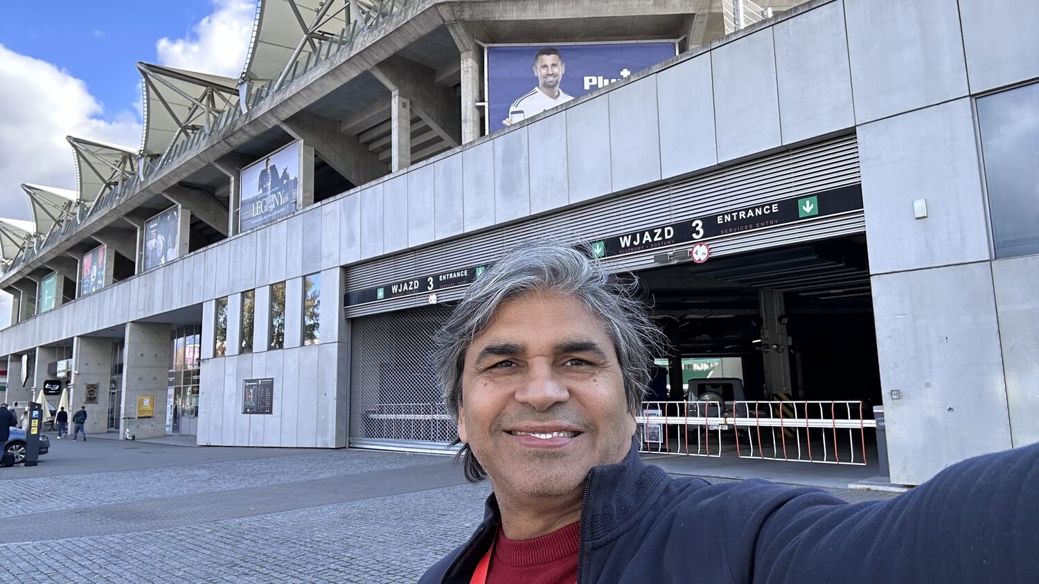 Tarry Singh outside the stadium at Wjazd 3, the concrete stand and Legia banners overhead under a bright autumn sky.