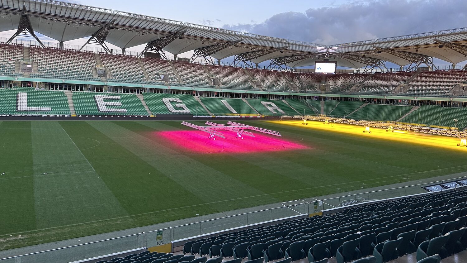 The view from the conference terrace over Legia Warsaw's pitch, the seat mosaic spelling LEGIA across the far stand, grow lamps burning pink and yellow on the turf.