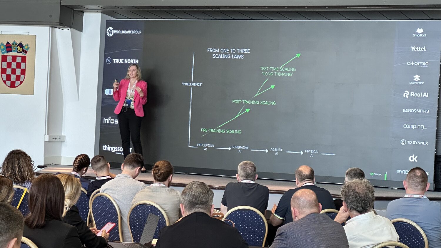 Delegates seated in rows watching a slide about scaling laws, the speaker on stage at the left.