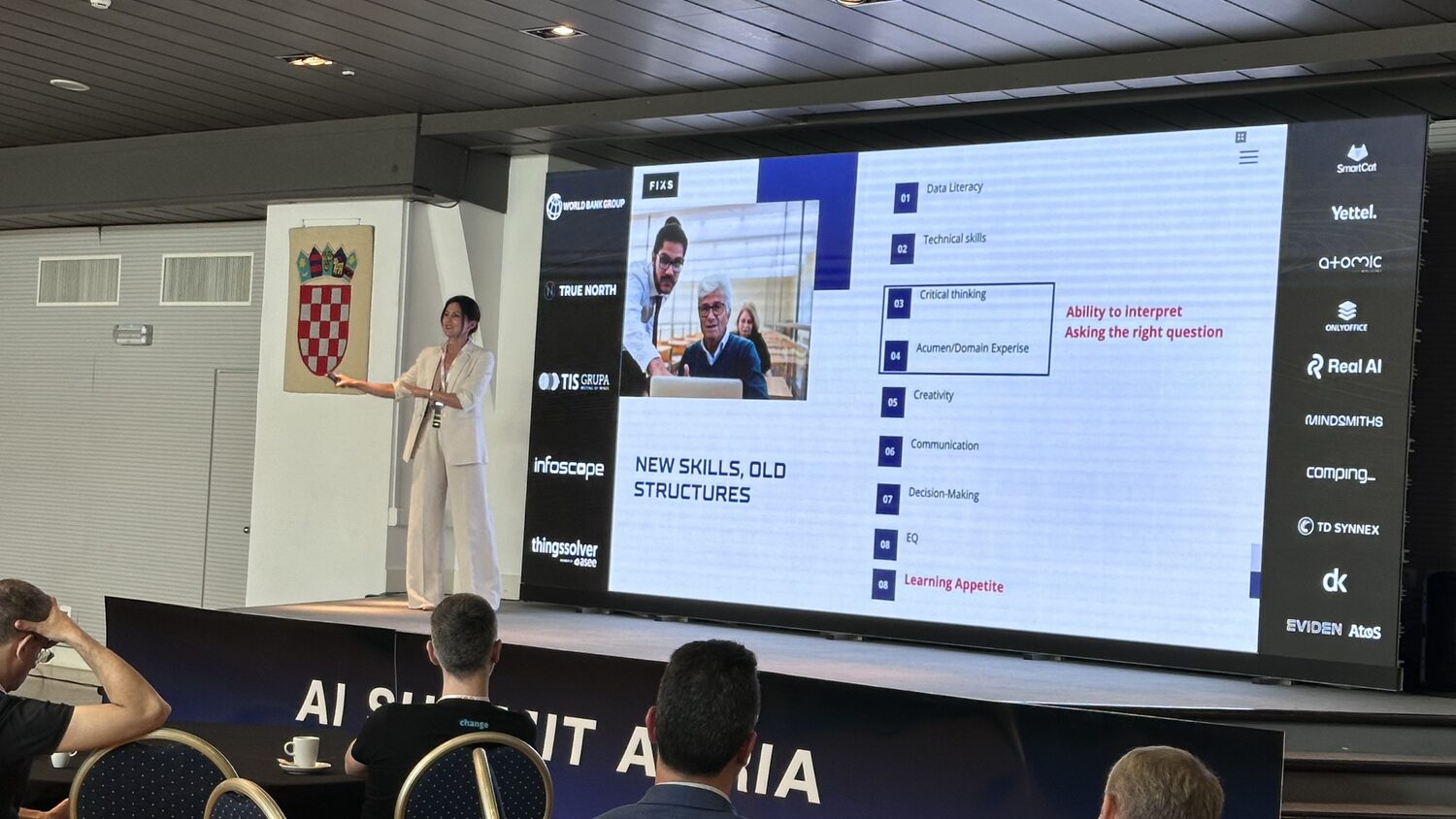A speaker on stage in front of a slide headed New Skills, Old Structures, with the AI Summit Adria banner along the stage front.